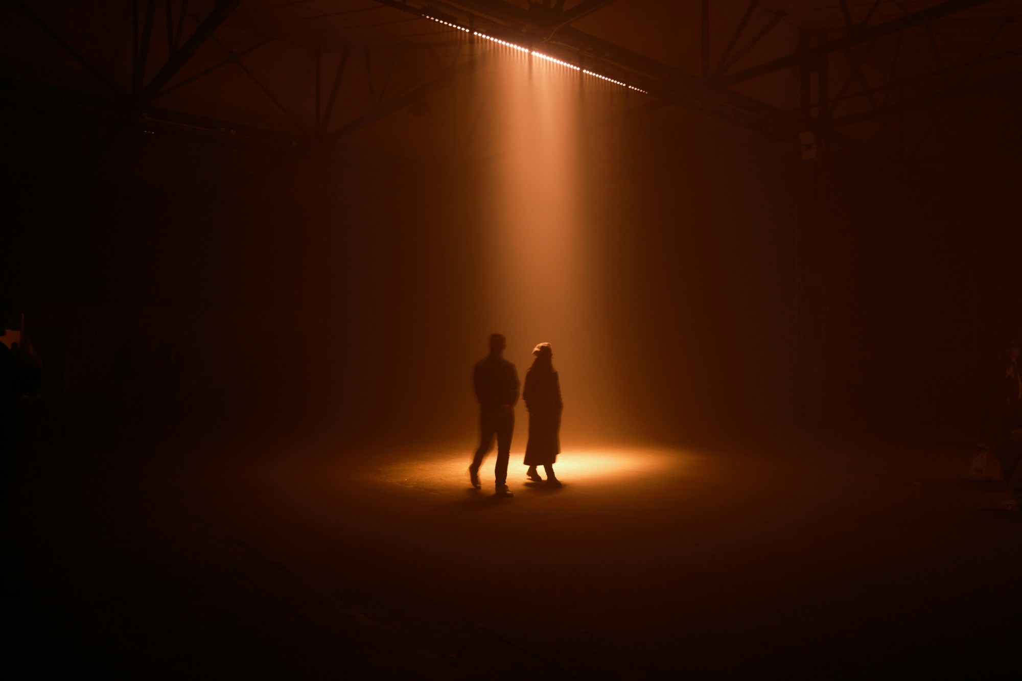 Two people standing in a single beam of warm light in a dark warehouse